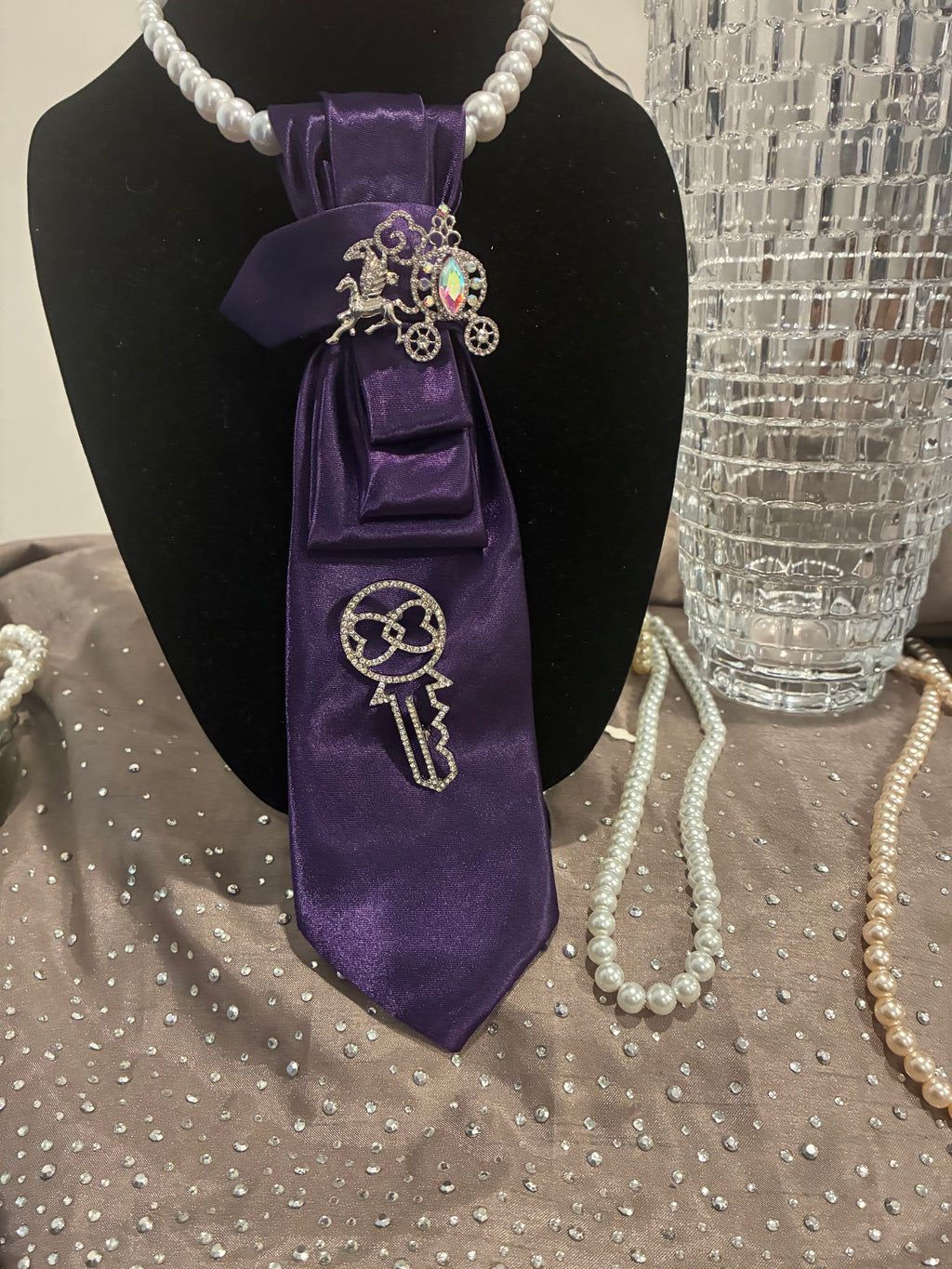 A Purple satin necktie with a rhinestone key broach and horse drawn carriage broach and pearl necklace
